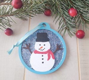 Round wooden hanging ornament with snowman in the foreground and blue-silver glitter background.